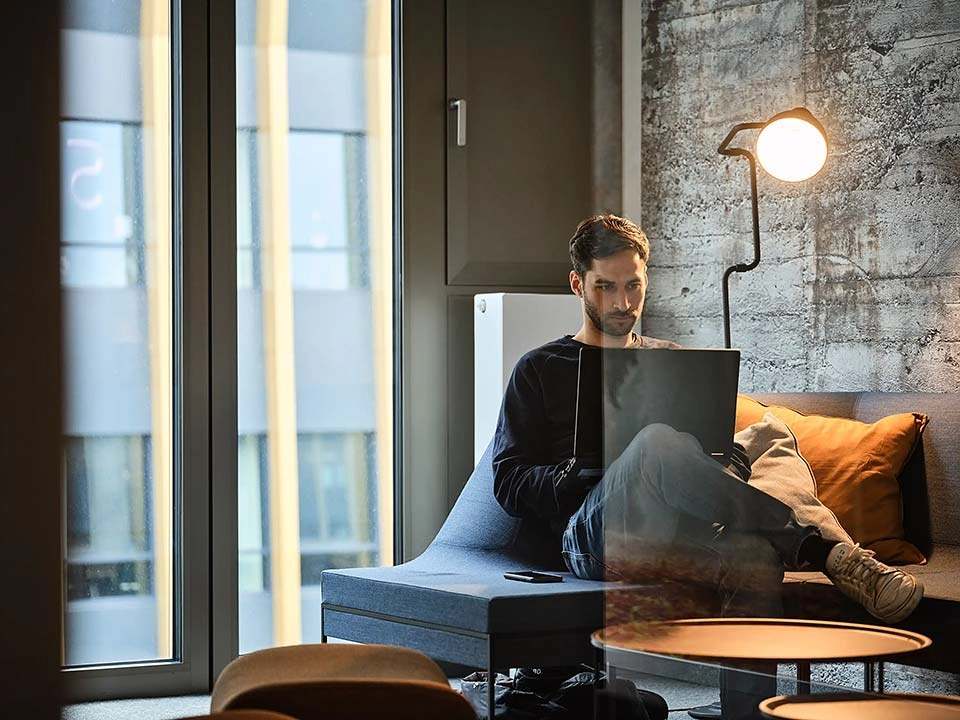 Ein Mann sitzt mit überschlagenen Beinen auf einer Couch im isolierten Büro und arbeitet am Laptop